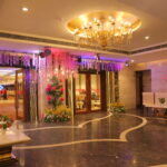 Elegant banquet hall entrance with floral decorations, golden chandelier, marble flooring, and seating area leading to the main event space.