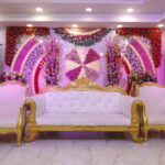 Luxurious wedding stage with white and gold ornate sofa set, vibrant floral wall backdrop, and decorative lighting arrangements.